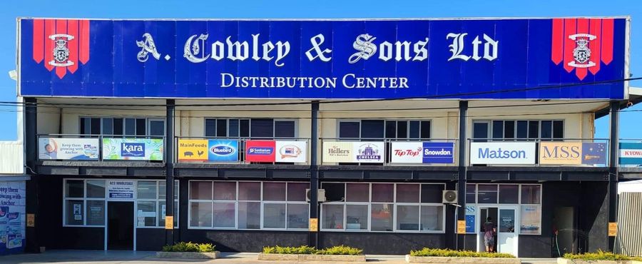 The A. Cowley & Sons Ltd Distribution Center in Nuku'alofa, its blue facade carrying the company name in blackletter above a row of brand boards including MainFeeds, Bluebird, Sanitarium, AFFCO, Hellers, Chelsea, Tip Top, Snowdon and Matson.