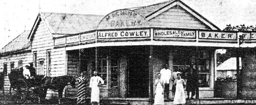 Alfred Cowley's bakery in Nuku'alofa: a timber shopfront with a deep verandah and a painted sign reading Alfred Cowley, Wholesale and Family Baker, with staff in white aprons on the step and a horse and cart loading at the side.