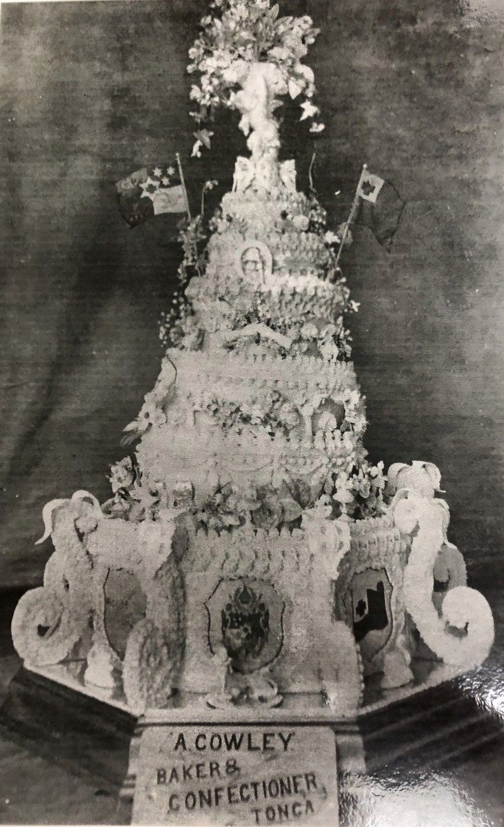 The rebuilt wedding cake standing in tiers, decorated with piped scrollwork, floral sprays, the flags of Tonga and New Zealand, and armorial panels, with a card at its base reading A. Cowley, Baker and Confectioner, Tonga.