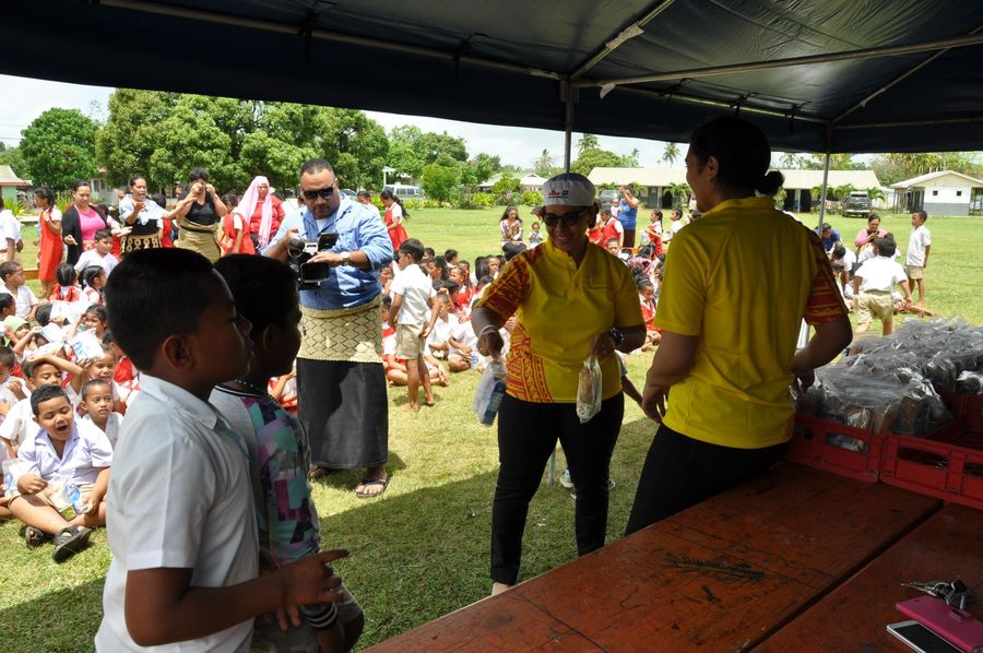 ACS volunteers in yellow shirts handing packed lunches to students under the marquee, with a cameraman filming behind them.