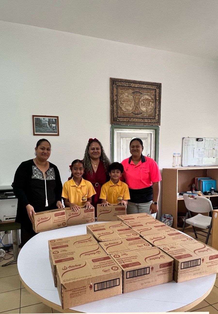 Ita and Joseph in their yellow school uniforms with school staff, behind a table stacked with donated cartons of Anchor chocolate milk.