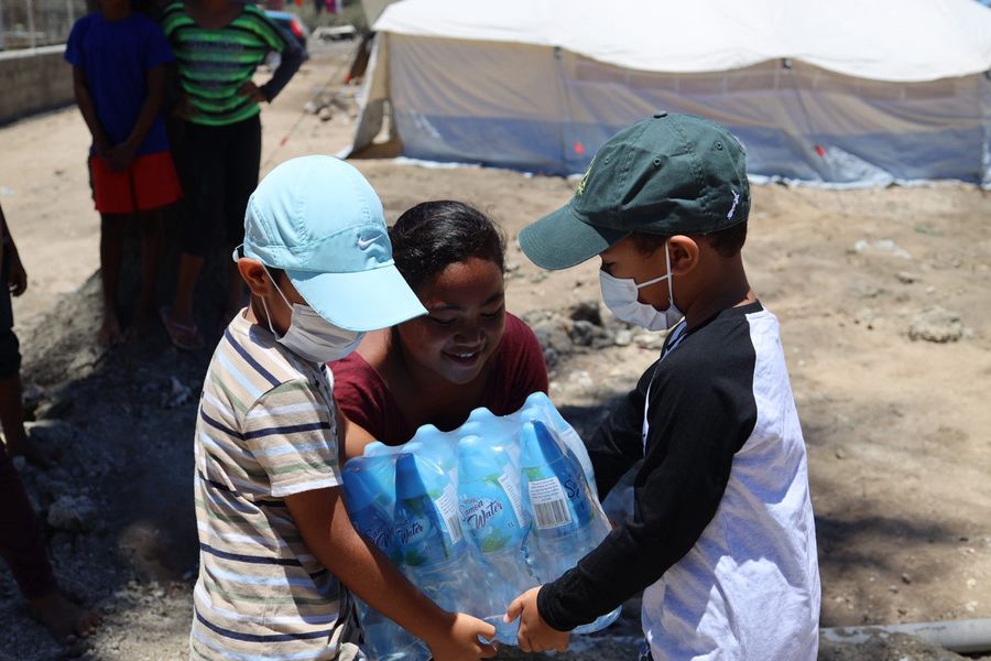 Alfred and Buk, in caps and face masks, handing a pack of bottled water to a smiling child beside the relief tents.