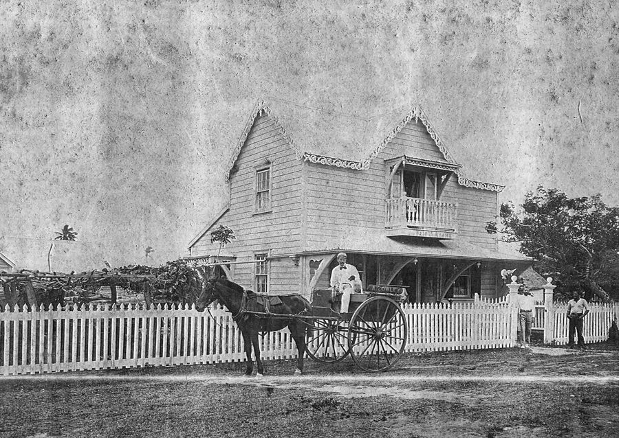 A two-storey timber house with decorative bargeboards behind a white picket fence, a horse and gig waiting at the gate with A. Cowley painted on the side panel, and a man in baker's whites and cap holding the reins with a dog beside him.