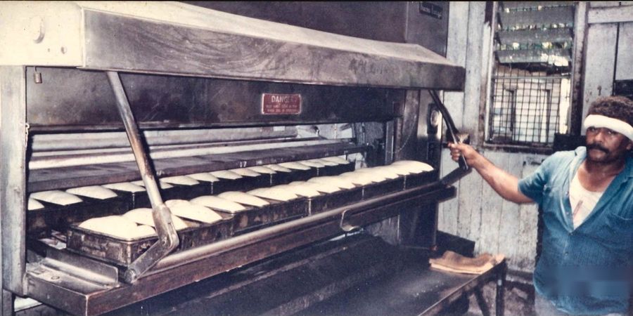 A baker drawing a long tray of tinned loaves from the oven.