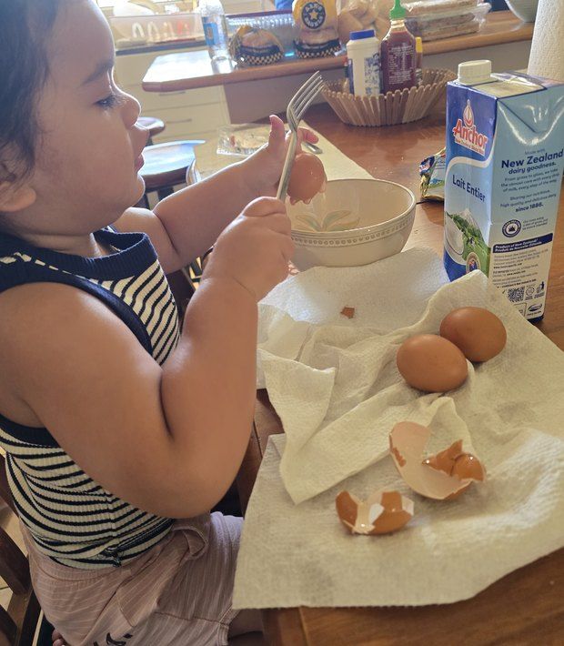 A toddler cracking eggs into a bowl at the kitchen bench.