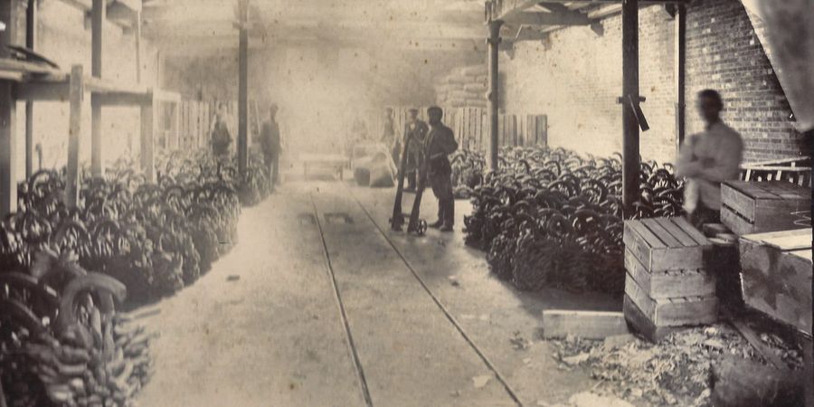 A produce shed with banana bunches stacked either side of a rail line, wooden crates and workers with a hand trolley.